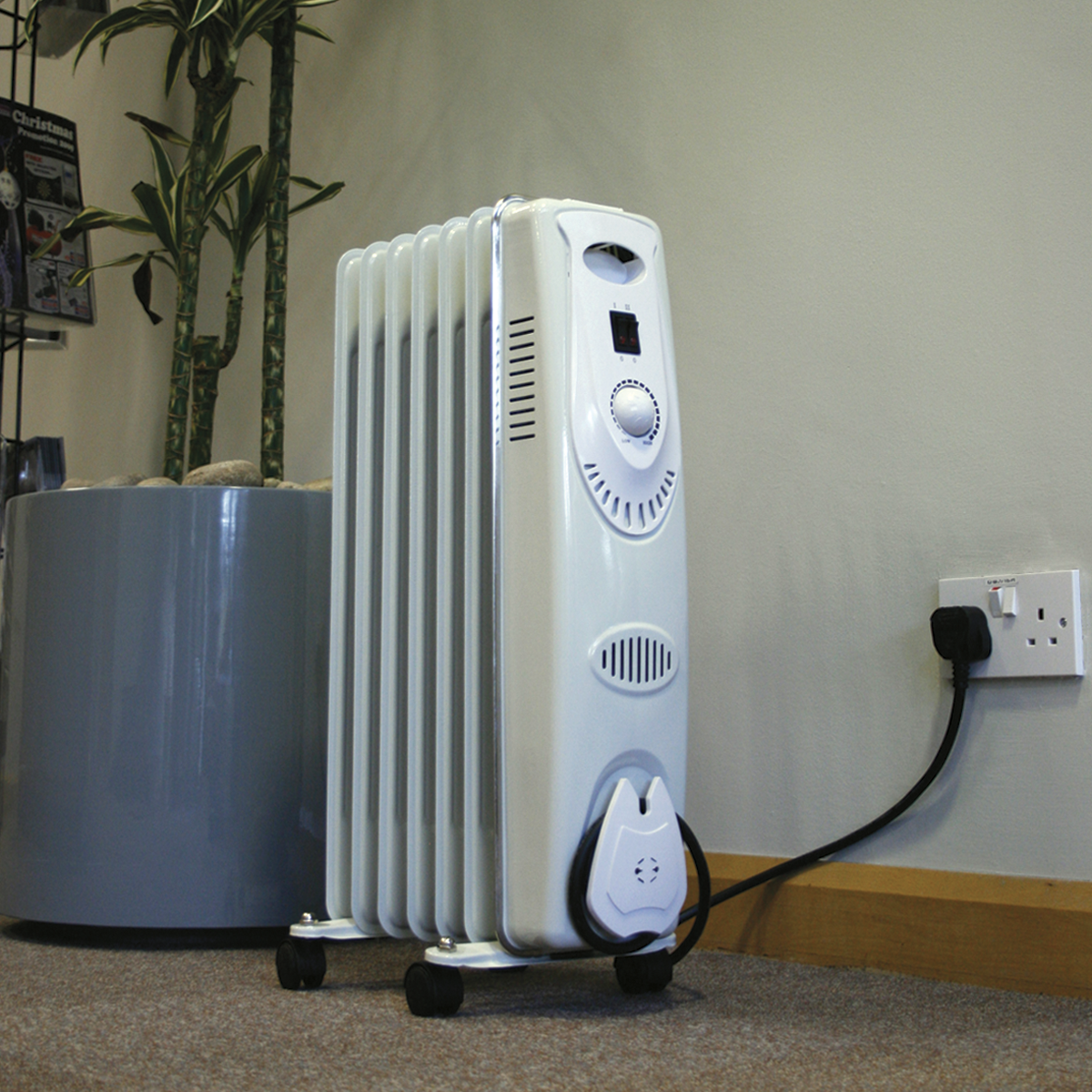 A Sealey Oil Filled Radiator 1500W/230V 7-Element - RD1500, featuring a safety tip-over switch and thermostatic heat control, is plugged into a wall socket in a room with a plant in a large gray pot.