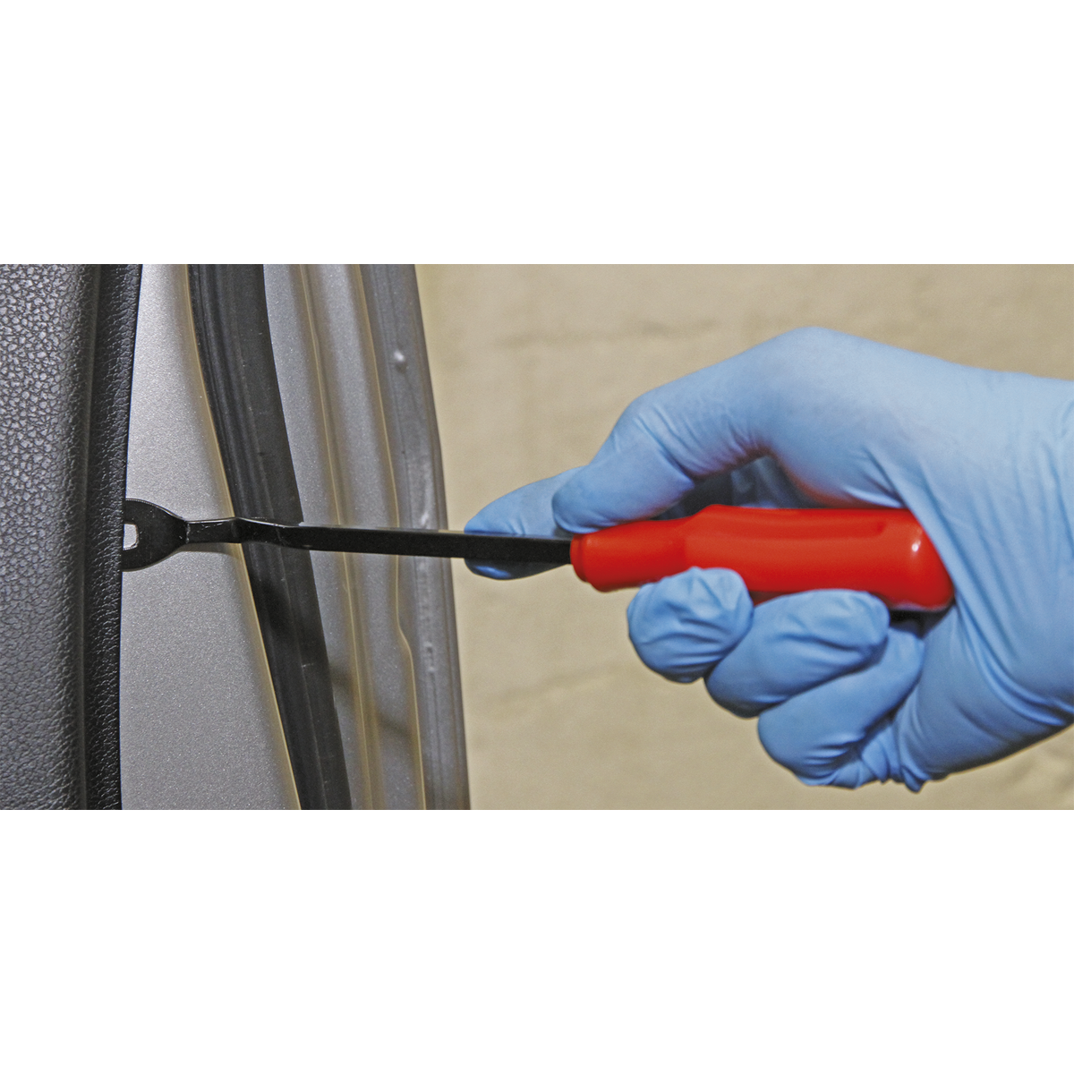 A gloved hand uses the Sealey Trim Clip Tool 220mm - RT008 with a red handle to pry trim clips from a car door panel.