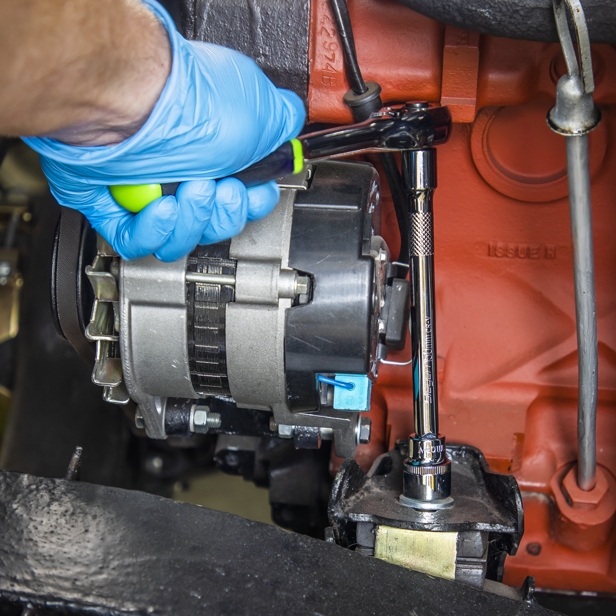 A person wearing blue gloves uses a 72-Tooth ratchet wrench to adjust an alternator component in an engine. Nearby, the Sealey Socket Set 12pc 3/8"Sq Drive WallDrive® Metric (S01242), made of durable Chrome Vanadium steel, is neatly organized and ready for use.