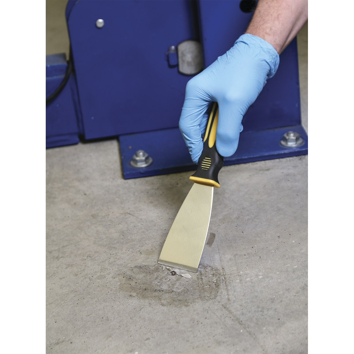 A person wearing a blue glove utilizes the Sealey Rigid Blade Scraper Set 3pc - S0600 to remove a substance from a concrete floor adjacent to a blue metal structure.