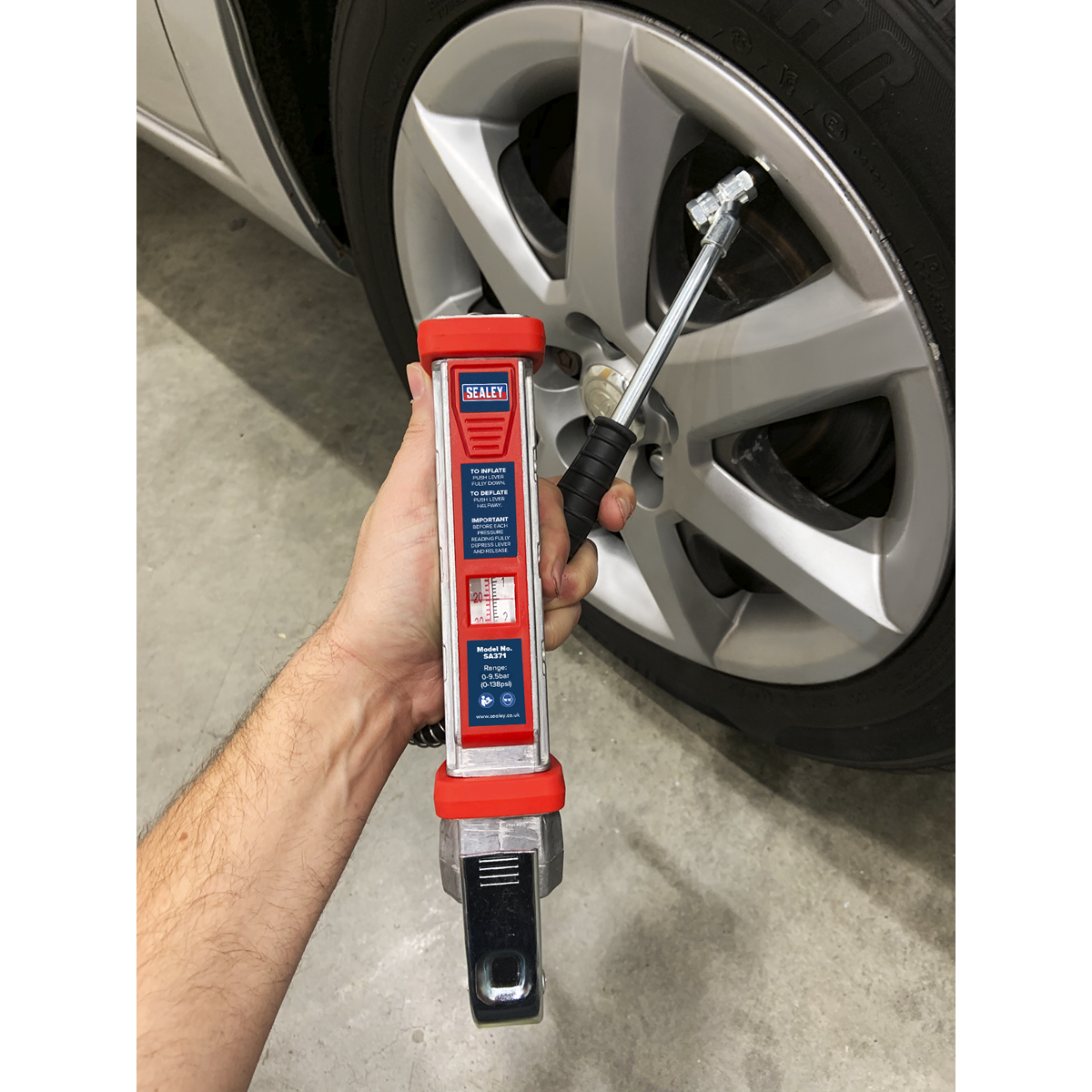 A hand holding a Sealey Tyre Inflator 0.5m Hose with Twin Push-On Connector (Model SA371) with a tire pressure indicator, positioned in front of a car wheel. The aluminium body ensures durability and precision, making it an essential tool for any vehicle.