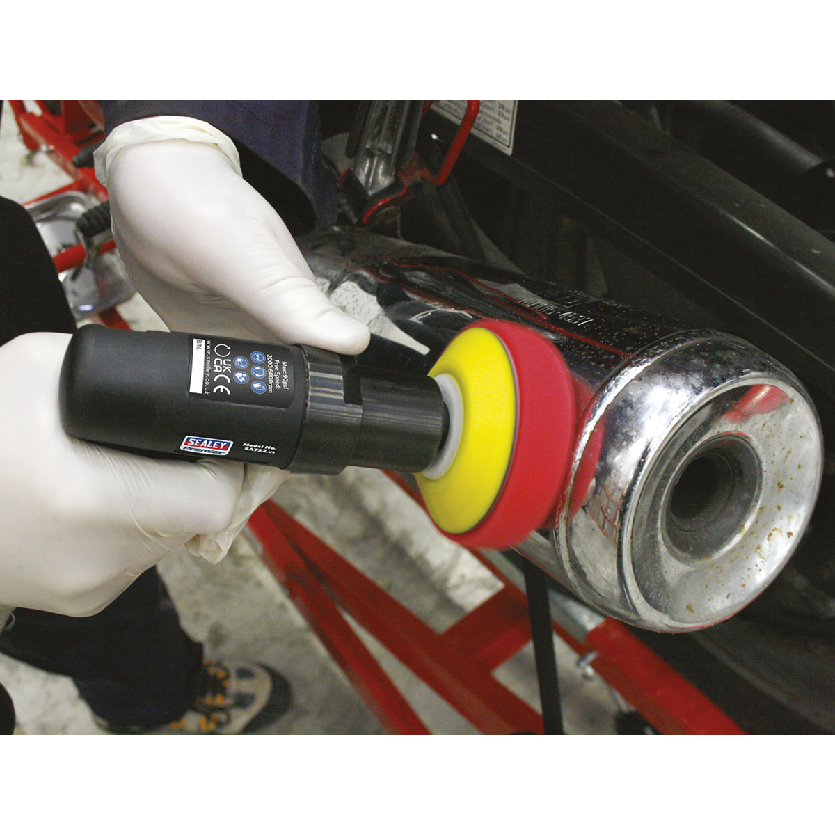 A person wearing gloves uses the Sealey Premier Composite Mini Air Polisher Ø75mm - SA722, with its spinning red and yellow buffer pad, to polish a metal surface while benefiting from its adjustable speed control.