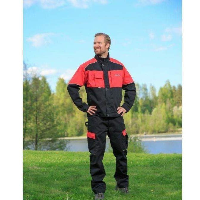 A person wearing a durable Valtra - Work Jacket - V4280510 in black and red from the AGCO brand stands on a grassy field with trees and a lake in the background. The safety-focused attire includes reflective material for visibility. The "Massey Tractor Parts" logo is displayed at the bottom left.