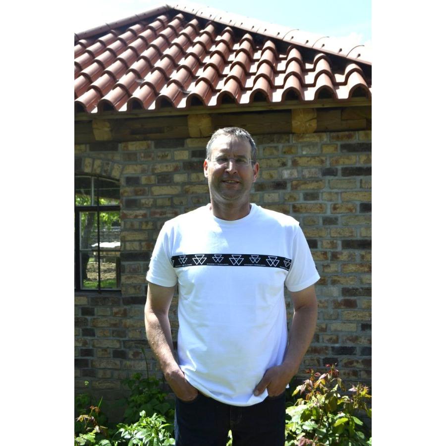 A man wearing an AGCO Massey Ferguson White Crew Neck T-Shirt (Product Code: X993412302000) stands outside in front of a brick building with a tiled roof.