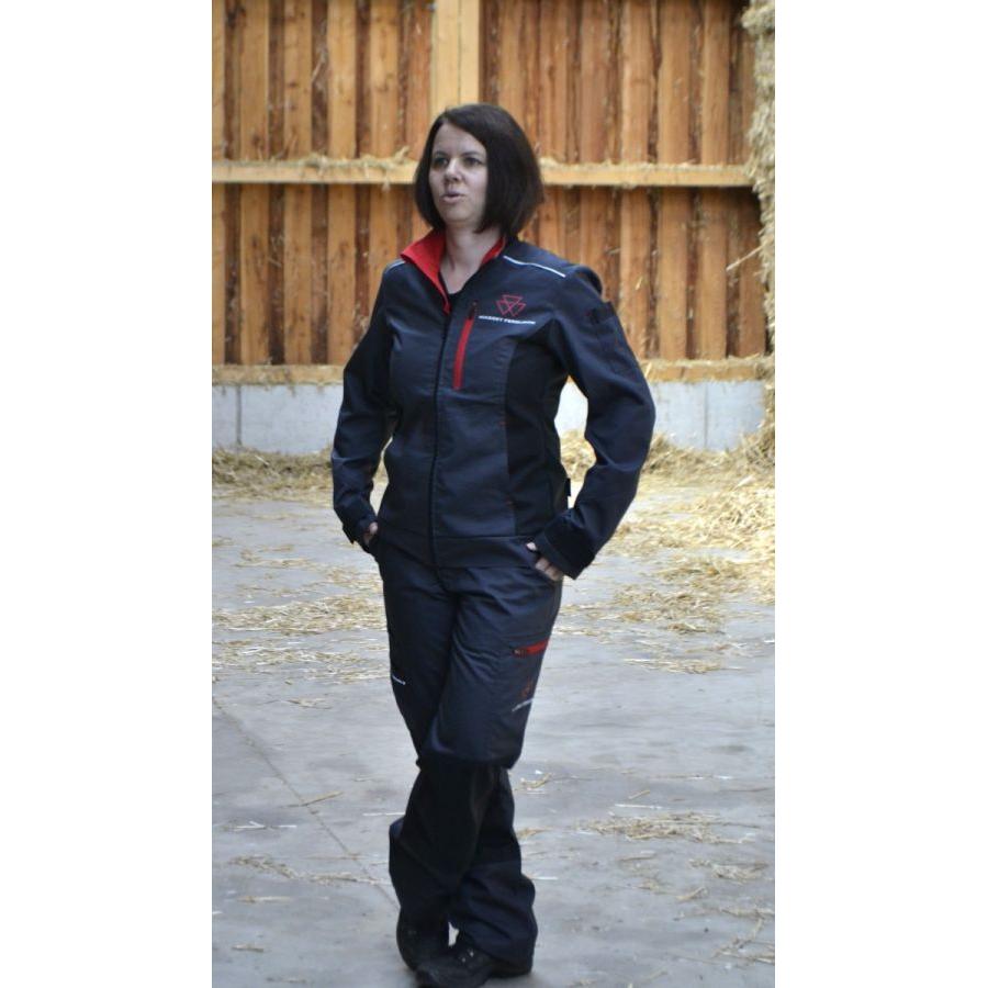 A person wearing the AGCO Massey Ferguson Women's Work Jacket (X993532214), featuring adjustable cuffs and reflective stripes, stands on a concrete floor in a barn with hay scattered in the background.