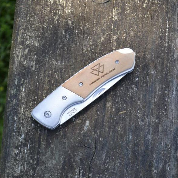 An AGCO Massey Ferguson folding pocket knife, model X993562301000, with a stainless steel blade and a metallic brown handle showcasing a geometric design, placed on a wooden surface.