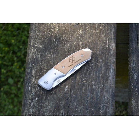 An AGCO Massey Ferguson folding pocket knife, model X993562301000, with a stainless steel blade and a metallic brown handle showcasing a geometric design, placed on a wooden surface.