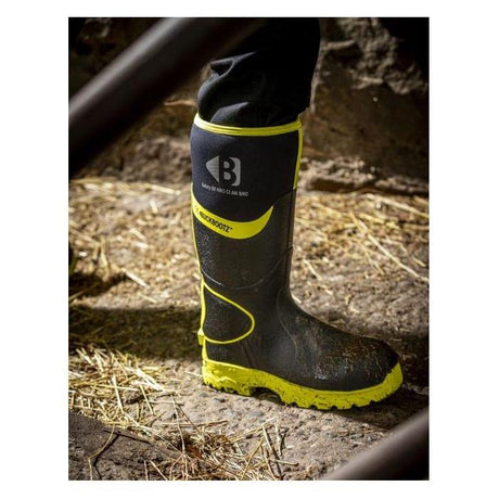 A person wearing JMCE Buckler Hi Viz Safety Wellies in black and yellow stands on dirt and hay. The boot, marked with the letter 'B' and the words "superbootz," features lightweight components known as BBZ8000 for enhanced comfort.