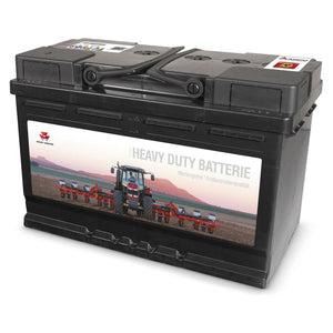 The AGCO Massey Ferguson Batterie, 72 Ah - X991450325000 features a heavy-duty design with a label showcasing an image of a tractor on a farm at sunset. This battery incorporates Calcium-Technologie for enhanced performance and has prominent top terminals along with branded markings in the top corners.