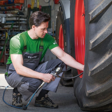 A man in a green and gray work uniform crouches beside a large tractor tire, using a pneumatic tool to inflate the tire. The setting appears to be a workshop or farm garage with equipment in the background.