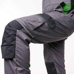 Close-up of dark gray work pants with black knee panels, green contrast stitching, and a bright green inner lining visible at the leg opening, shown on a cropped model against a white studio background.
