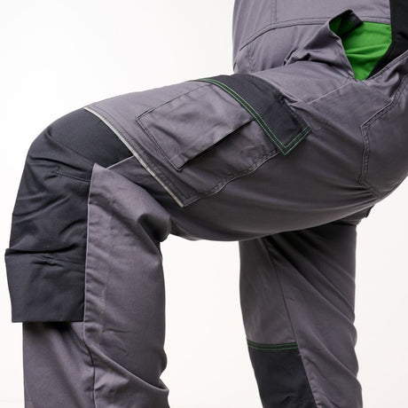 Close-up of dark gray work pants with black knee panels, green contrast stitching, and a bright green inner lining visible at the leg opening, shown on a cropped model against a white studio background.