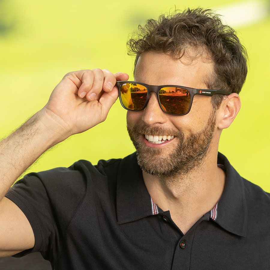 Man with a beard, wearing a black polo shirt and AGCO Massey Ferguson sunglasses (model X993472201000), smiles as he lifts one arm to adjust his UV protection glasses. The background is a bright, blurred green.