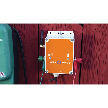 A Luda Fence Alarm (S.156395) from Sparex is mounted on a red wooden surface with a green object partially visible on the left side. The orange device, complete with wires and indicator lights, ensures reliable GSM coverage for seamless operation with the FenceAlarm app.