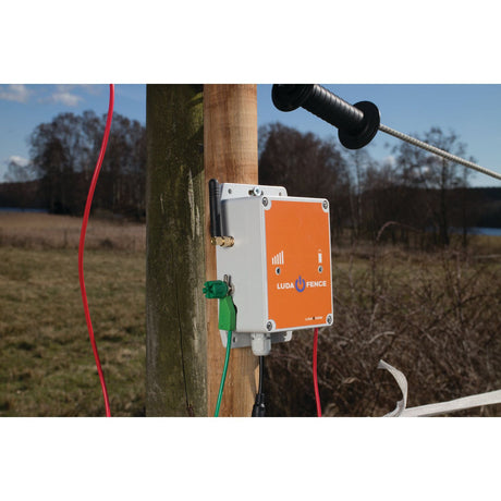 A Fence Alarm - Luda - S.156395 by Sparex is installed on a wooden post in an outdoor, rural setting with fields and trees in the background, guaranteeing reliable GSM coverage and seamless integration with the FenceAlarm app.