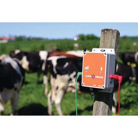 A Sparex Fence Alarm - Luda - S.156395 control device is attached to a wooden post in a pasture with black-and-white cows in the background, monitored through the FenceAlarm app.