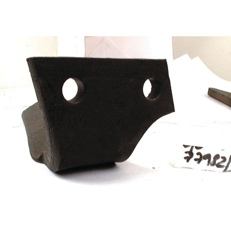 A black, irregularly-shaped metal bracket with two circular holes, each 17mm in diameter and spaced 68mm apart, is placed on a white surface. Handwritten text on a white piece of paper is partially visible on the right side, possibly indicating its compatibility as a replacement for Kuhn machinery. This part appears to be the Sparex Power Harrow Blade 110x14x350mm RH (52552600 | Sparex Part No.S.77982).