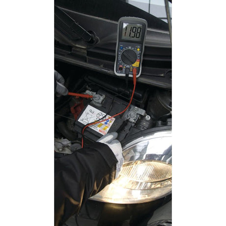 A person wearing gloves uses the Sparex Digital Multimeter - S.151762 to check the Voltage DC of a car battery, which displays a reading of 11.98 volts.