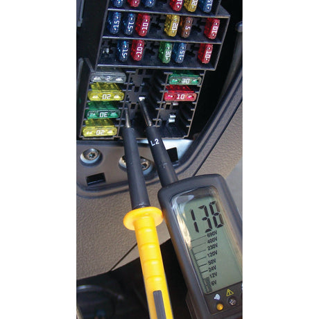 An Electrical Voltage tester - S.151758 from Sparex is being used to measure voltage at a car's fuse box. The display shows 13.5 volts.