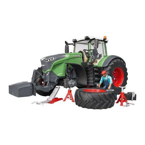A mechanic wearing a red cap works on the Fendt 1050 Vario tractor, distinguished by its adjustable rear coupling, currently elevated on red jacks. Scattered around are various pieces of workshop equipment and a detached tire. This setup is part of the AGCO's product range under the model X991016003000.