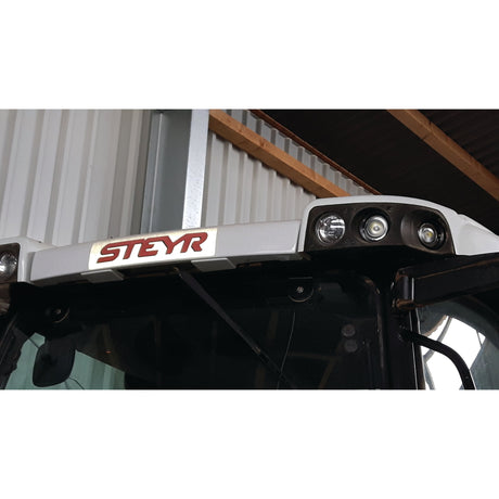 Front view of a Steyr tractor featuring its LED Work Light (Cree High Power) from Sparex with 3000 lumens raw brightness and a roof logo. The background includes a corrugated metal wall and part of a wooden roof structure.