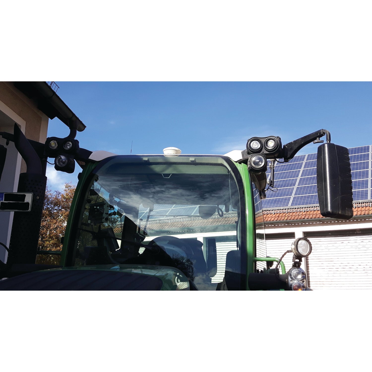 A close-up of a green tractor's cabin featuring multiple lights and mirrors, including the Sparex LED Work Light (Cree High Power) with an integrated 2 pin Deutsch connector, parked near buildings with solar panels on the roof.
