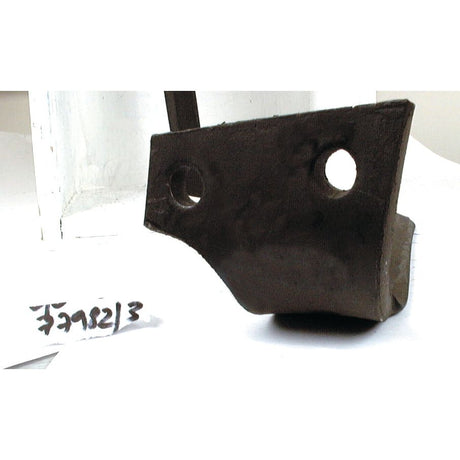 Close-up of a dark, irregularly shaped metal object with two holes, identified as a Sparex Power Harrow Blade 110x14x350mm LH, placed next to a piece of white paper with writing on it. This replacement blade for Kuhn features hole centers at 68mm and a hole diameter of 17mm, designed to fit model number 52552500 and carries the Sparex Part No. S.77983.