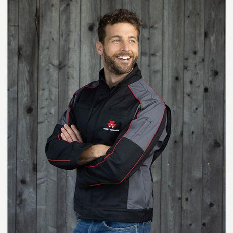 A man with a beard smiles, wearing an AGCO Massey Ferguson S Collection Work Jacket (X993482105) in durable black and gray with a red and white logo, standing in front of a wooden wall.