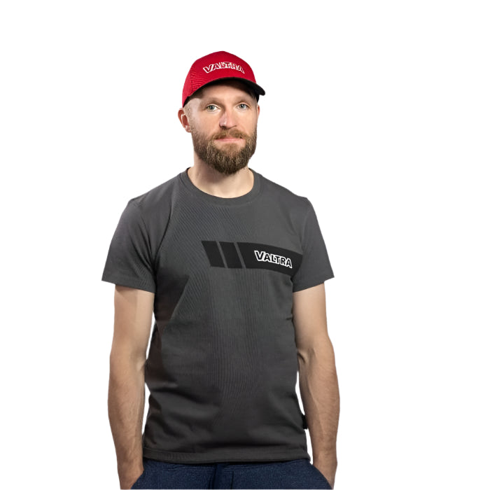 A bearded man is wearing a gray VALTRA t-shirt made of 100% cotton and a red Valtra cap (model: V42501406) by AGCO with a slide buckle, standing against a plain white background.