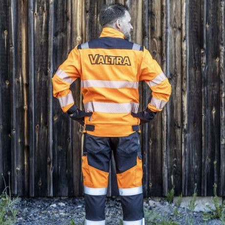 Person facing away, wearing AGCO's Valtra High Visibility Work Trousers (V428093) in orange and black, crafted from durable four-way stretch material with "VALTRA" printed on the back. They stand in front of a wooden wall, and the trousers feature practical cargo pockets for easy storage.