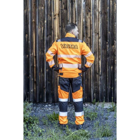 Person facing away, wearing AGCO's Valtra High Visibility Work Trousers (V428093) in orange and black, crafted from durable four-way stretch material with "VALTRA" printed on the back. They stand in front of a wooden wall, and the trousers feature practical cargo pockets for easy storage.